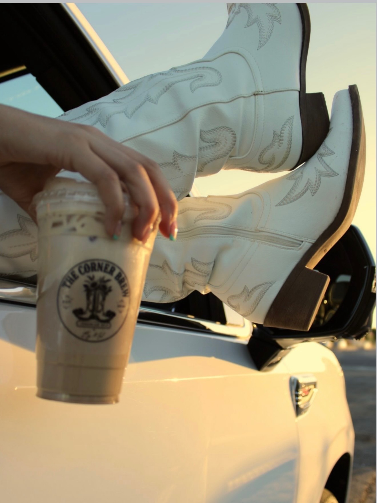 White cowboy boots out a car window at sunset with a hand holding an iced Corner Brew coffee.